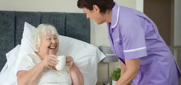 elderly lady in bed with hot drink female carer purple uniform 2x1 1 768x359 1.webp